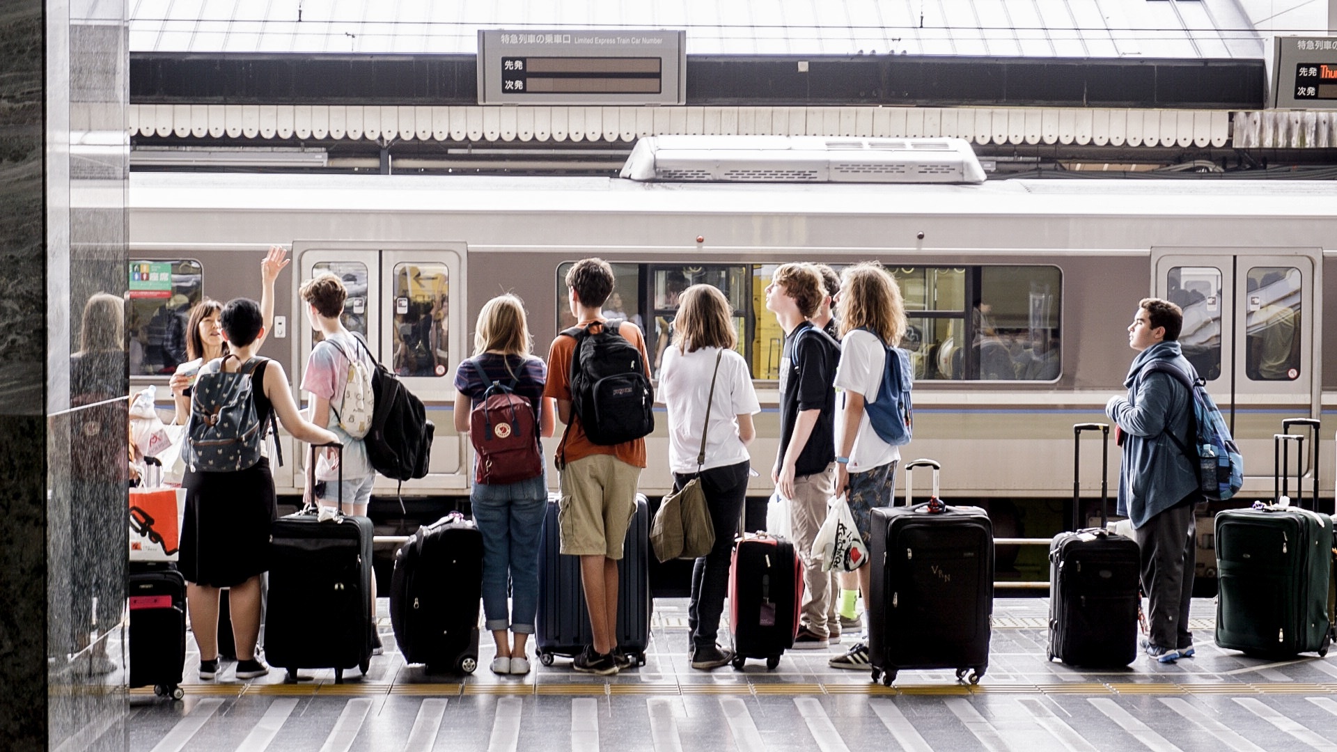 A group of travelers with suitcases and backpacks stand on a train platform facing a stopped train. The indoor station features signage in Japanese and English, indicating an international travel setting.