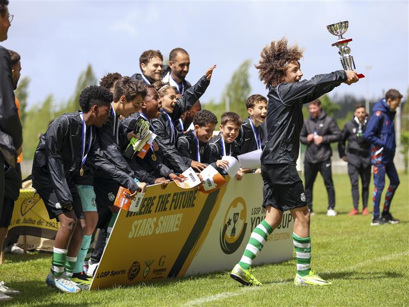 Young soccer players celebrate a victory on a grassy field, with one player holding up a trophy and others wearing medals. A banner in front reads 'WHERE THE FUTURE STARS SHINE!' alongside sponsor logos, capturing the excitement of a youth sports competition.