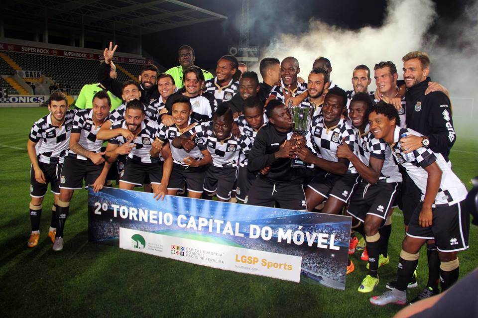 Soccer players in black and white checkered jerseys celebrate a tournament win at night, holding a trophy in front of a banner reading '2º TORNEIO CAPITAL DO MÓVEL' with sponsor logos. The festive scene includes stadium lighting and background smoke or fog.