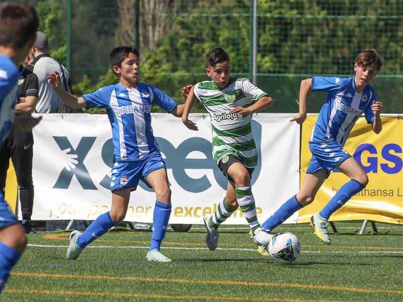 Three youth soccer players compete on an outdoor artificial turf field. One player in a green and white striped jersey dribbles the ball while two players in blue and white jerseys pursue. Advertising banners and greenery are visible in the background.
