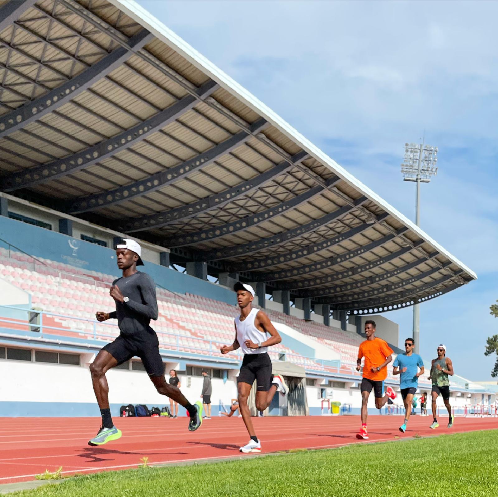 Six individuals run on a red track in an outdoor stadium with a large covered seating area in the background. They wear athletic gear, and the sunny weather with scattered clouds suggests a training or competition setting.