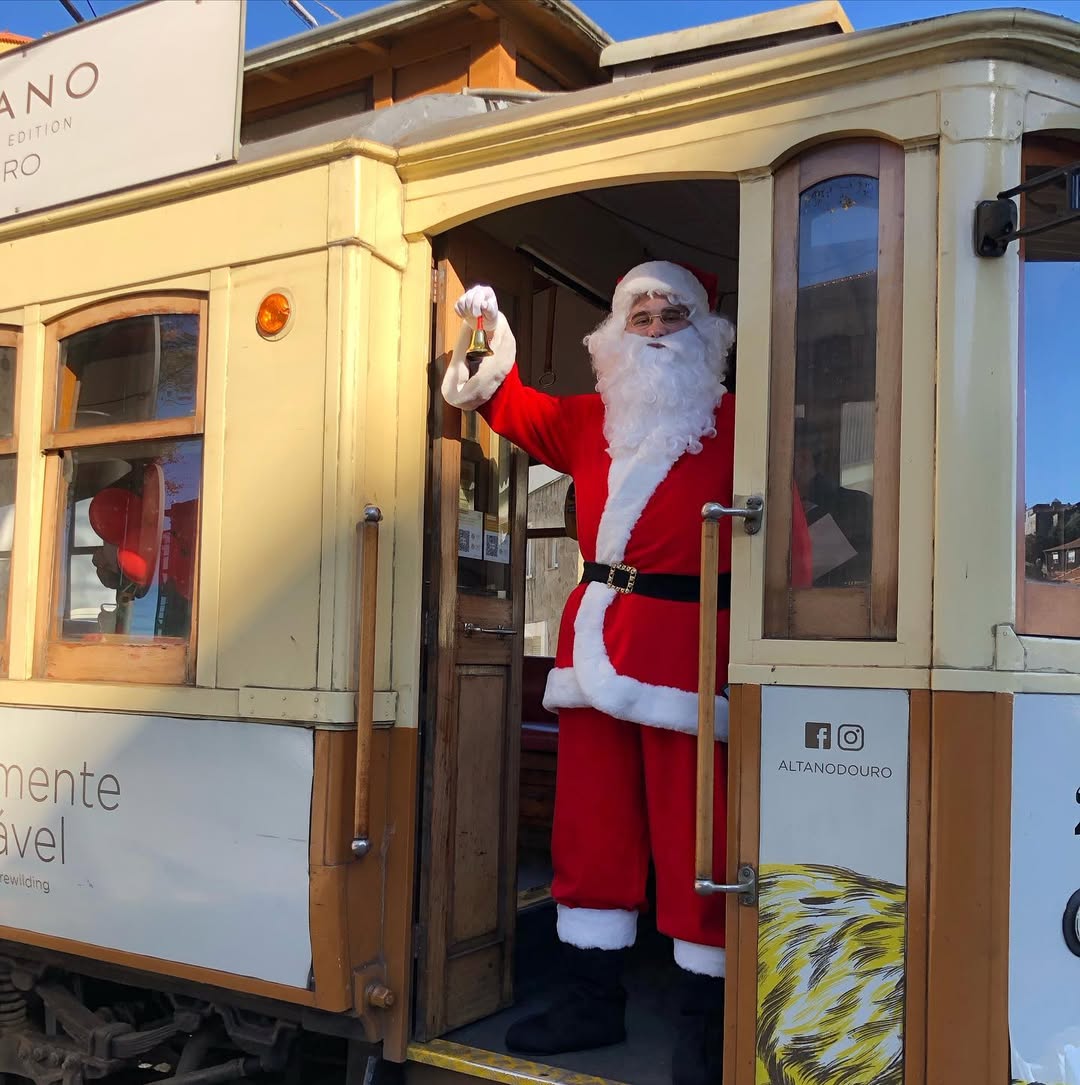 A person dressed as Santa Claus stands at the entrance of a vintage cream and yellow tram, holding a small bell. Signs on the tram include 'ALTANDOURO' and social media handles, creating a festive holiday scene with historic charm.